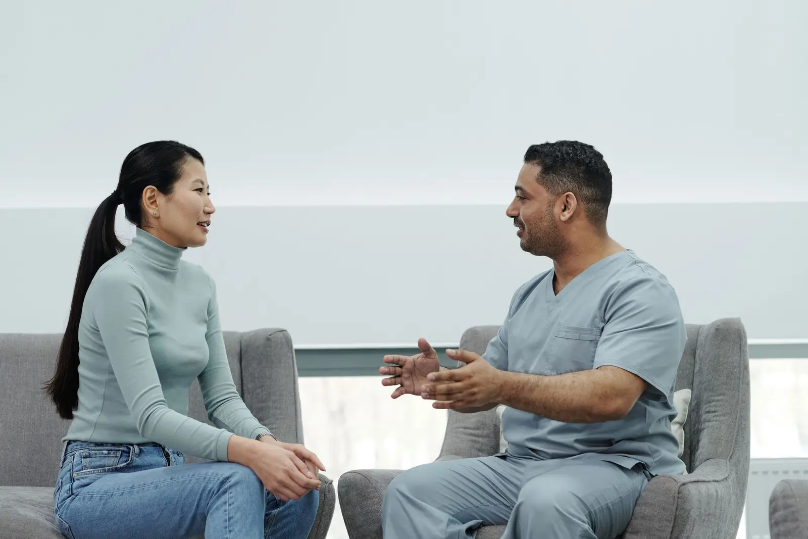 Clinician in scrubs talking with a patient as they sit facing each other in armchairs in a bright clinic